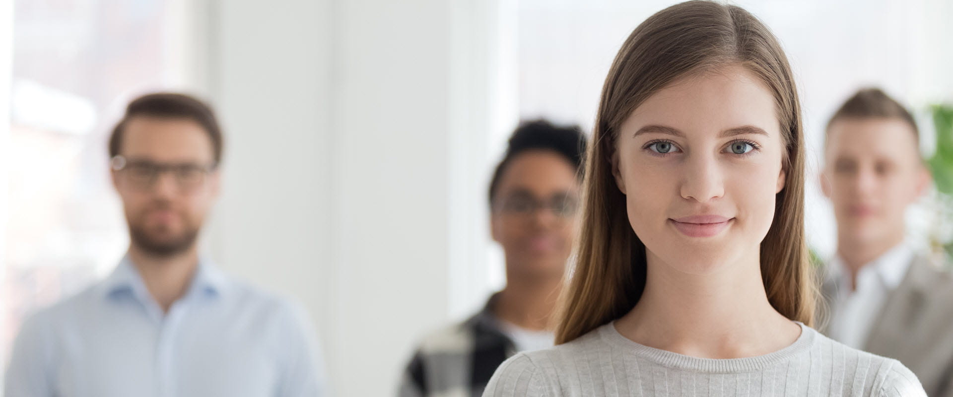 A confident young professional standing in front of their team members, who are slightly blurred in the background.